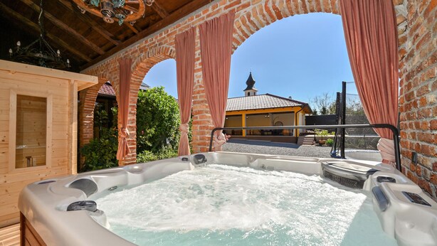 A hot tub area with a brick wall and wooden beams, a wooden cabin, and a building with a tiled roof in the background. - Cozy home in Gornja Ploscica (Gornja Ploscica)