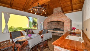 A rustic interior with a brick fireplace, wooden ceiling beams, wicker furniture, and a dining area with a view of greenery outside. - Cozy home in Gornja Ploscica (Gornja Ploscica)