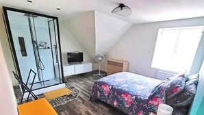 A modern bedroom with a glass shower, a TV on a white cabinet, a bed with floral bedding, and a small orange table.