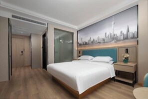Une chambre d’hôtel avec un grand lit, un tableau représentant un paysage urbain accroché au mur, une table de chevet avec un vase, et un plancher en bois.