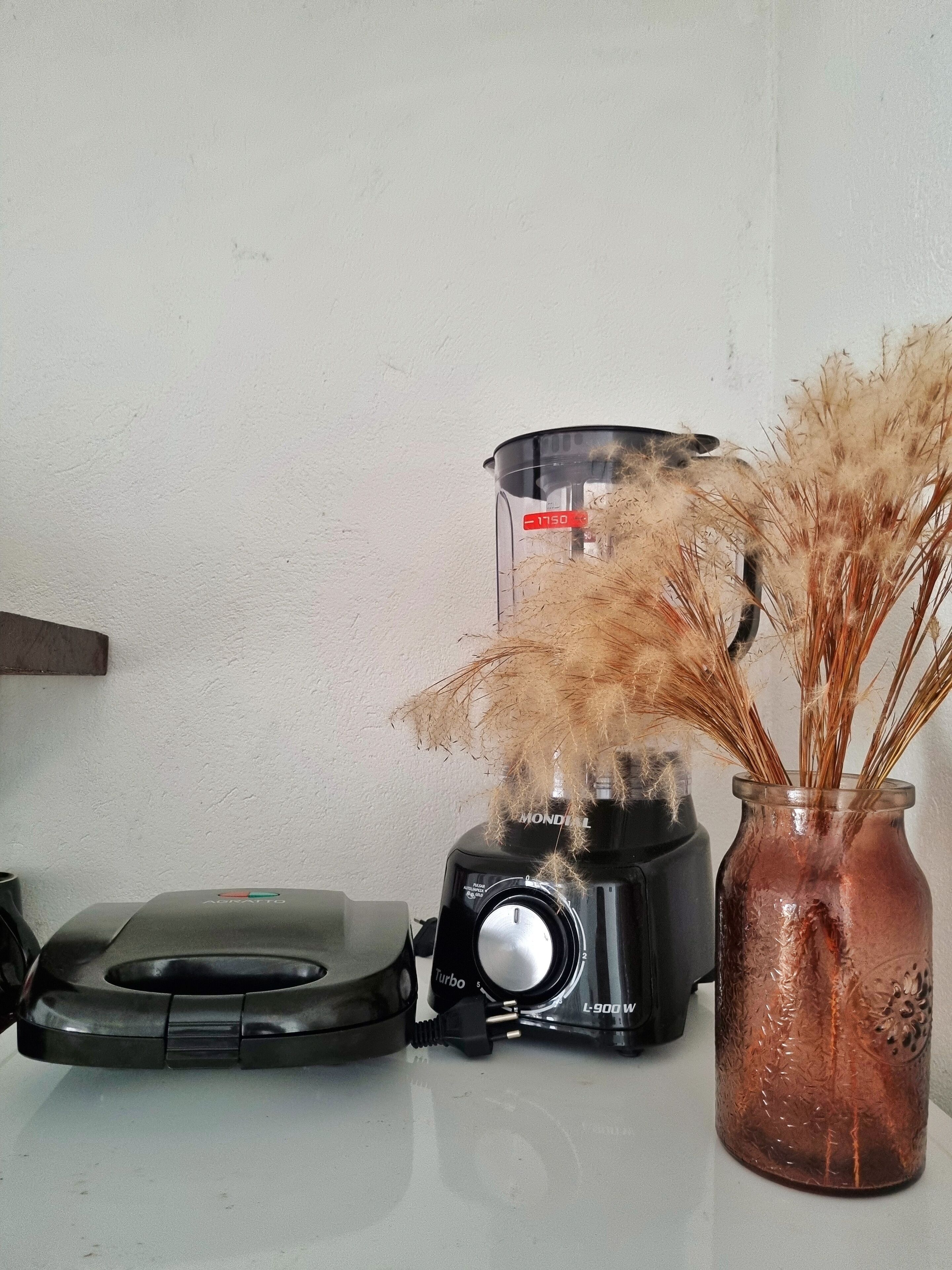A kitchen counter with a blender, a jar with dried plants, and a wall-mounted shelf.