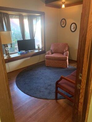 Una sala de estar con una televisión sobre una estantería de madera, un sillón rosa, un tapete azul y una puerta de madera.