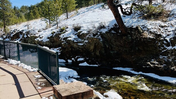 A snow-covered outdoor area with a concrete pathway, a metal railing, and a small stream. - Modern Luxury! Central Downtown Condo with parking! (License #6072) (Estes Park)