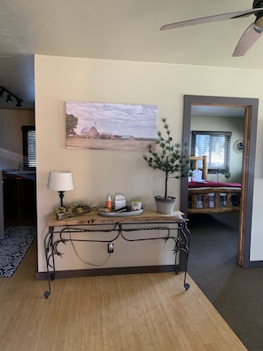 A room with a wooden floor, a metal console table with a lamp, a potted plant, and a painting of a rural landscape.