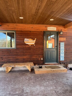 A wooden cabin with a dark green door, a sign reading 'Be Our Guest,' and a 'Home Sweet Home' sign.