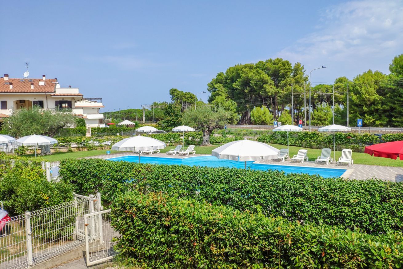 A pool area with lounge chairs and umbrellas, surrounded by greenery and a building in the background.