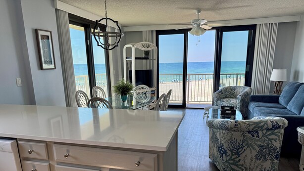 Une cuisine moderne dotée d’un grand îlot central, d’un coin repas avec des chaises blanches, d’un canapé bleu et d’une vue sur la plage grâce à de grandes fenêtres.