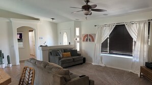 A living room with a grey sofa, a wicker chair, a ceiling fan, and a window with white curtains.