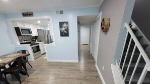 Una cocina moderna con armarios blancos, un microondas y un refrigerador. Hay una zona de comedor con una mesa y sillas. El piso es de madera y las paredes están pintadas de azul claro. Se ve una escalera a la derecha.