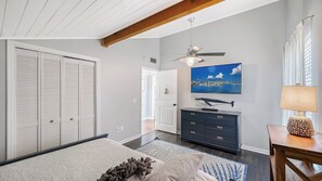 Une chambre à coucher comprenant un lit, une commode, un ventilateur de plafond, une télévision et une fenêtre avec des rideaux.