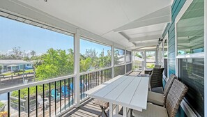 A covered balcony with a table and chairs, large windows, and a view of the surrounding area.
