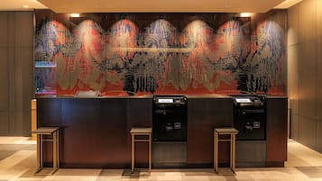 Un área de recepción con diseño moderno, que presenta un amplio mural abstracto en la pared, un mostrador de recepción y dos quioscos de autoservicio.