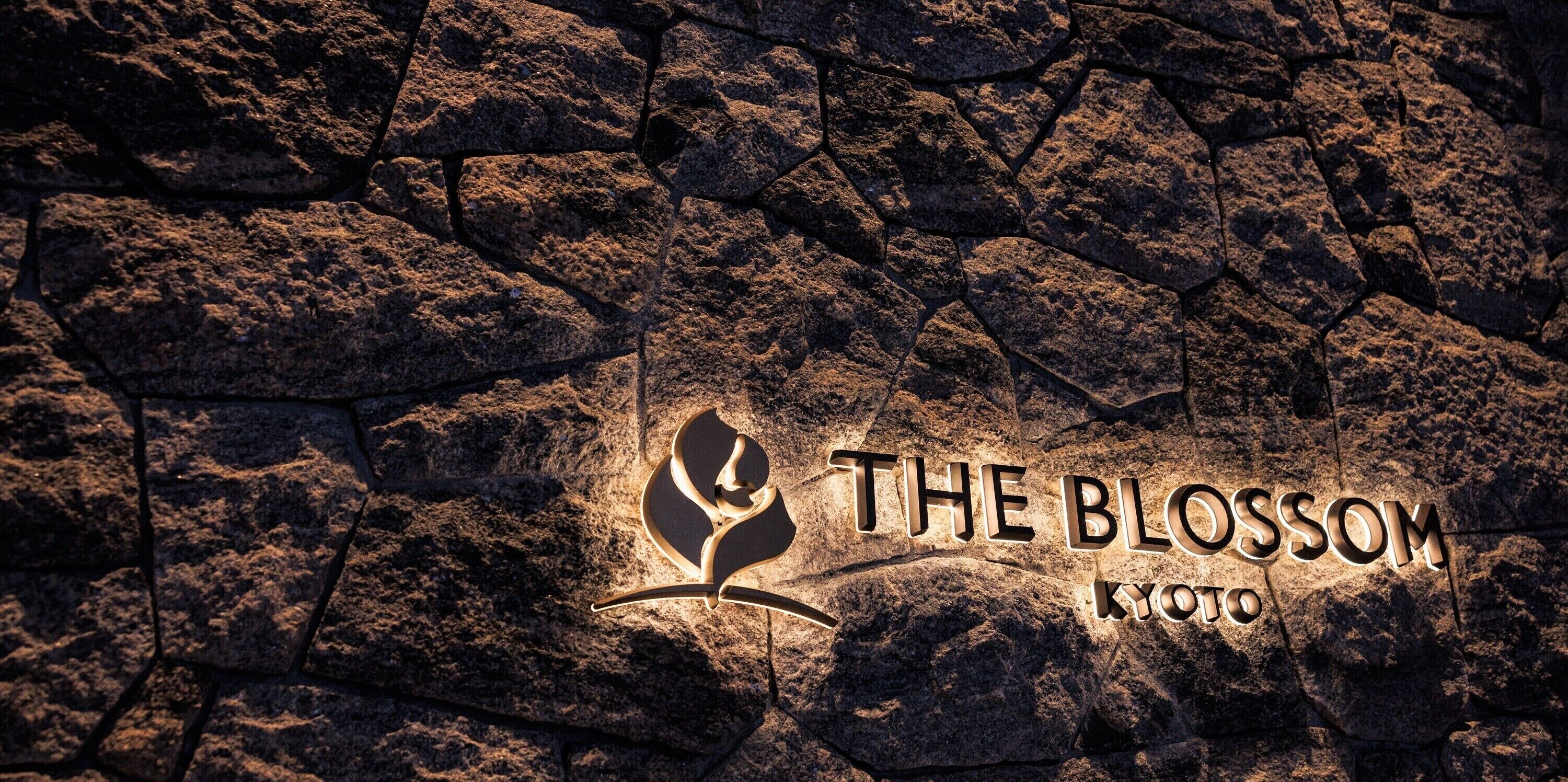 A logo with the text "THE BLOSSOM KYOTO" on a textured stone surface.
