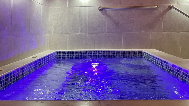 Une piscine intérieure moderne, dotée d’un éclairage bleu, de poutres en bois et de murs carrelés.