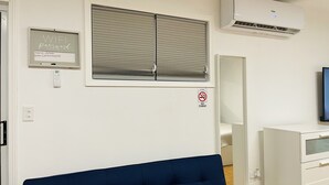 Une pièce avec un canapé bleu, une fenêtre avec des stores, un climatiseur, un téléviseur fixé au mur et un panneau indiquant qu’il est interdit de fumer.