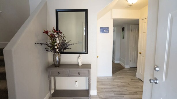 Un couloir avec une console grise, un miroir et un vase rempli de fleurs. Une porte se trouve sur la droite, et un autre couloir mène à une autre pièce.