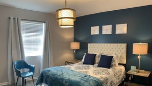 Une chambre à coucher comprenant un lit, deux tables de chevet, une chaise, une fenêtre avec des rideaux et un mur orné de trois tableaux.