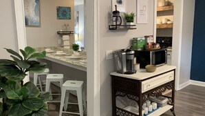 Une cuisine moderne avec des plans de travail blancs, une machine à café et un four à micro-ondes. On y trouve deux tabourets blancs et une plante en pot.