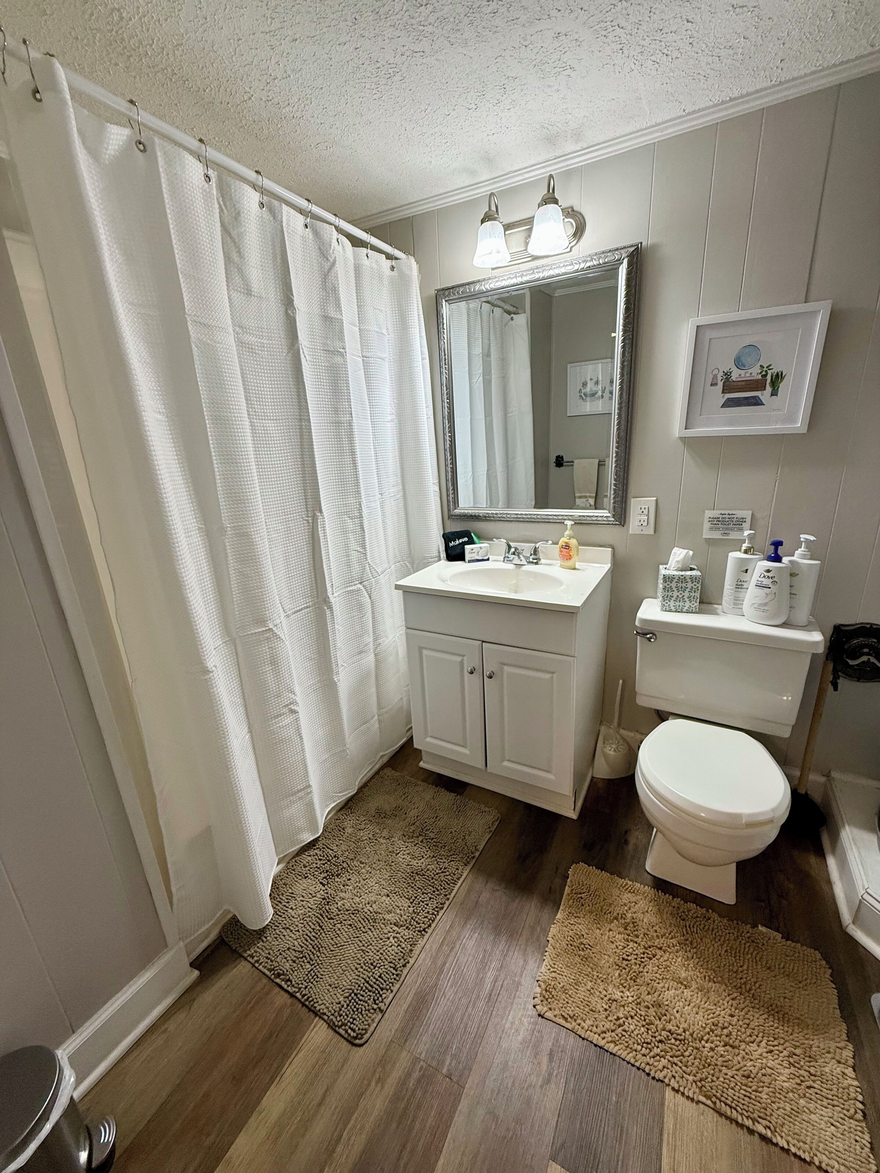 A bathroom with a shower curtain, a sink with a mirror, a toilet, and a rug on the floor.
