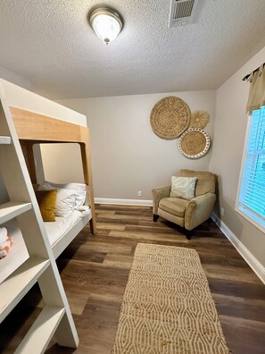 A room with a bunk bed, a chair, a woven wall decoration, and a patterned rug.