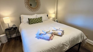Un lit bien fait, avec une couette blanche et des oreillers, un miroir rond accroché au mur, et une table de chevet sur laquelle se trouvent une lampe et une horloge.