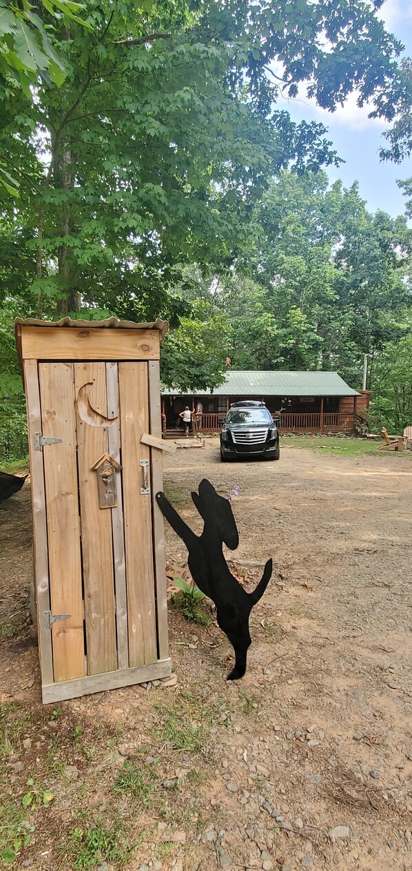 A rustic wooden structure with a metal door, a car parked in the background, and a person standing in front of it. - GUNSMOKE The not so "tiny"  cabin with fenced back yard! (Ellijay)