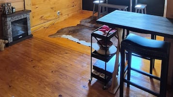 Interior de madera con chimenea, televisor, mesa de comedor y ventana con vista a follaje.