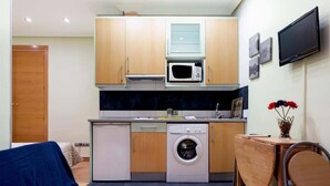 Een kleine keuken met een magnetron, oven en wasmachine, een eethoekje met een stoel, en een aan de muur bevestigde televisie.