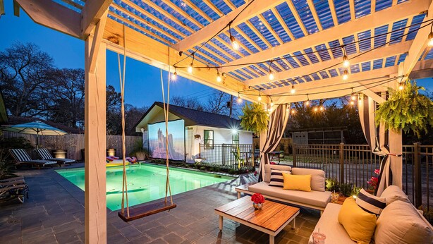 A covered outdoor patio with a swing, a pool, and a seating area with cushions and a table. - Charming Bungalow in Heights with pool/jacuzzi/game room. (Houston)