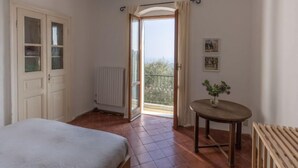 A bedroom with a bed, a radiator, a small table with a plant, and a balcony with a view.