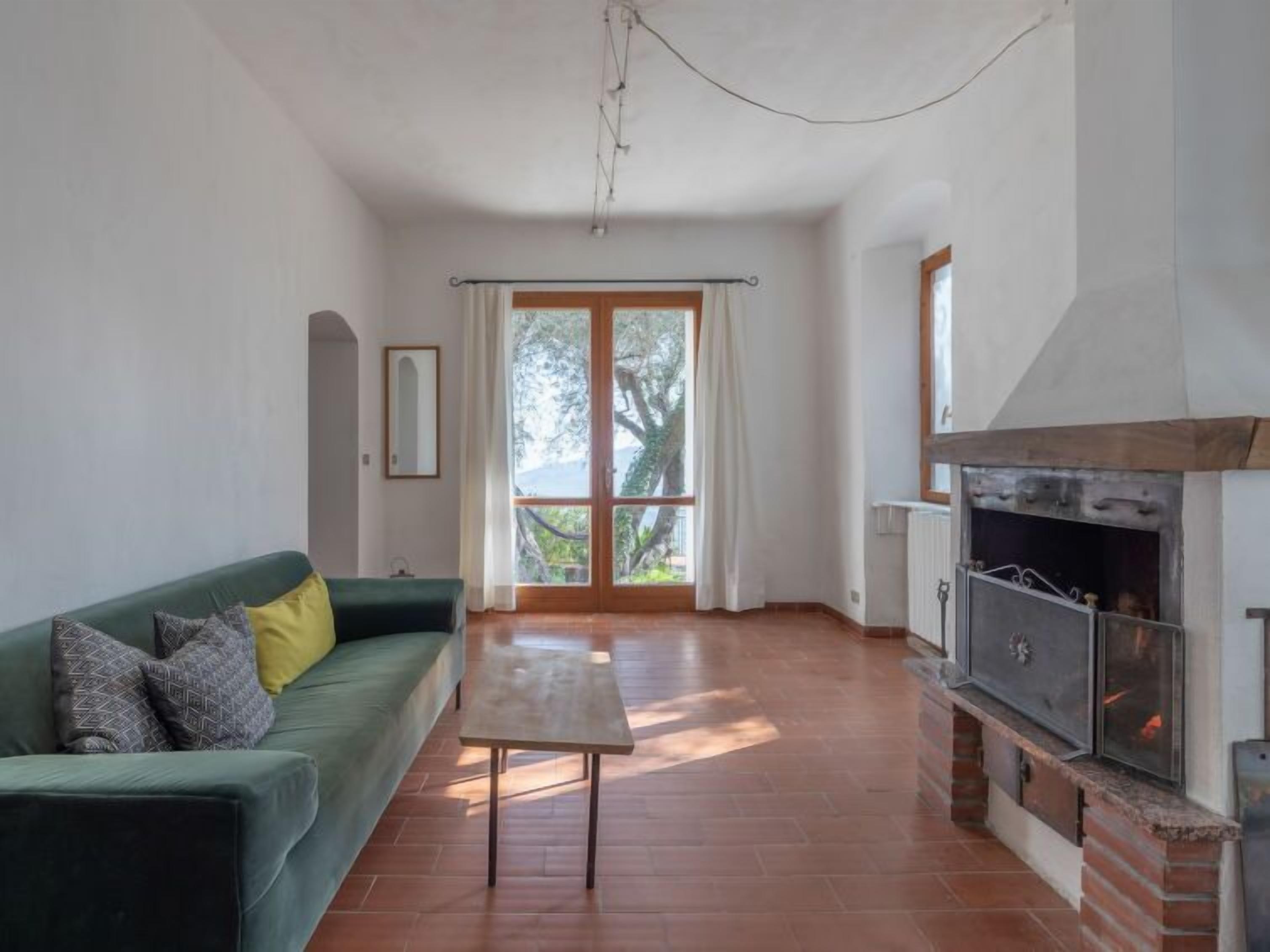 A living room with a green sofa, a small table, a fireplace, and a large window with curtains.