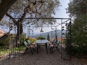 Un espace extérieur aménagé avec des sièges, protégé par un toit, comprenant une table et des chaises, entouré d’arbres et offrant une vue sur des collines lointaines.