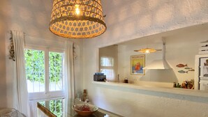 Un comedor con mesa de vidrio, lámpara colgante de mimbre, una silla blanca y una ventana con cortinas.