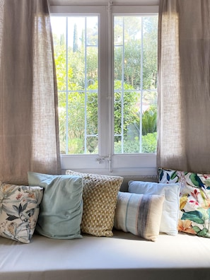 Una ventana con vistas a la vegetación, un marco blanco y un sofá con varios cojines.