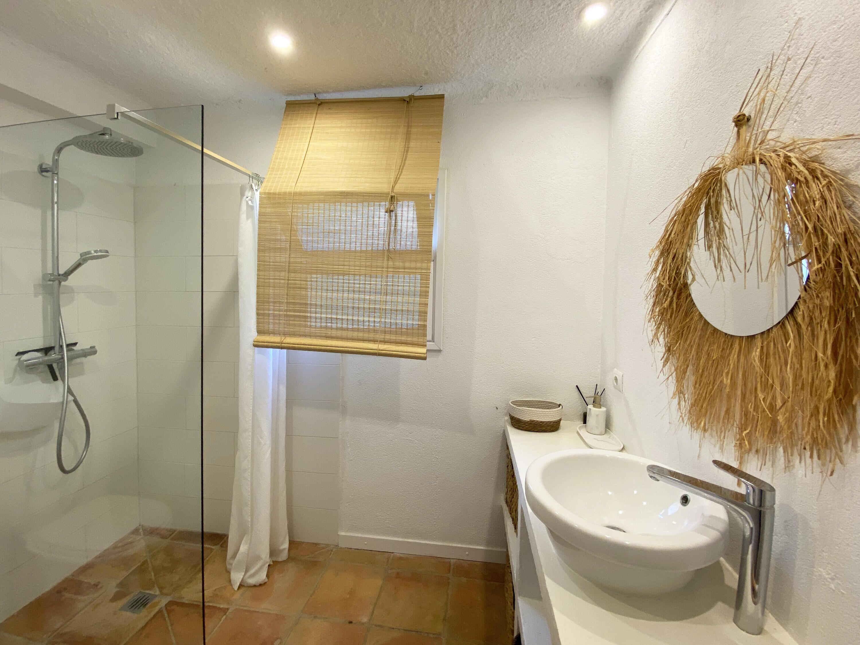 A bathroom with a glass shower enclosure, a white sink, and a round mirror with a straw-like decoration.