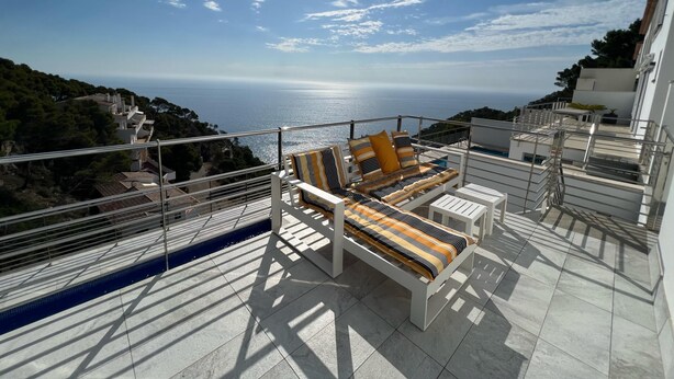 Um terraço com vista para o mar, com um sofá branco e almofadas listradas e uma piscina pequena.