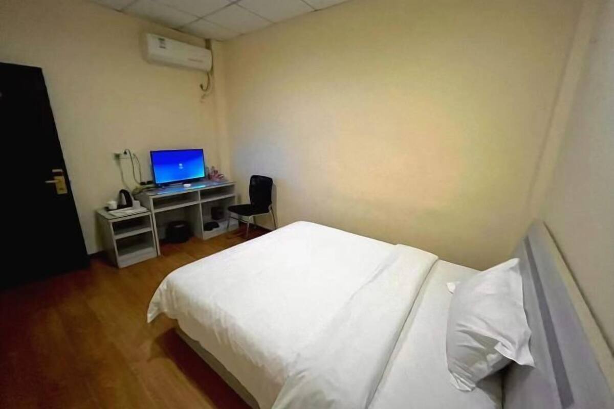 A hotel room with a bed, a desk with a computer, a chair, and a wall-mounted air conditioner.
