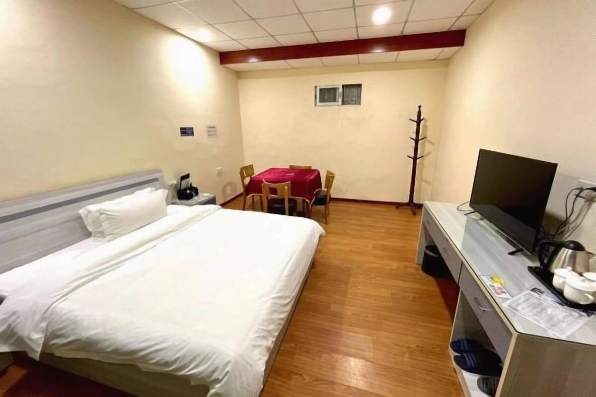 A hotel room with a bed, a desk with a TV, a chair, a small table, and a coat rack.