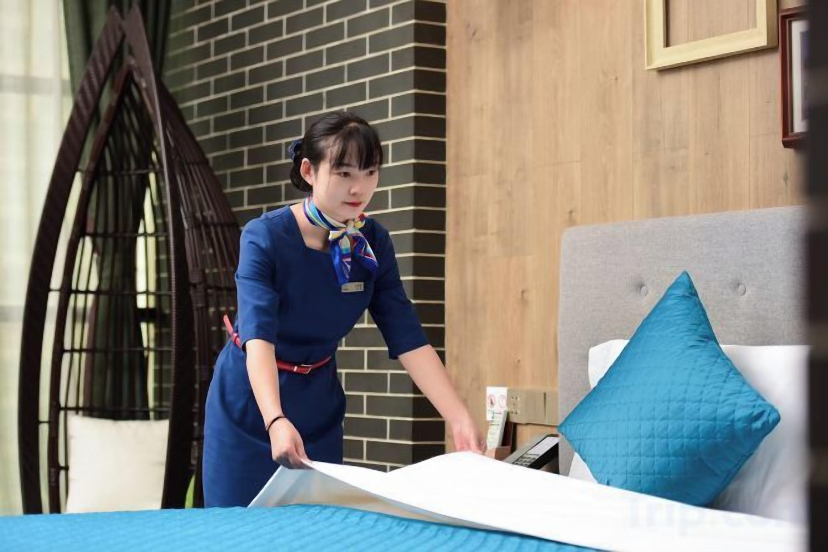 A person in a blue uniform cleaning a bed with a blue bedspread and a white pillow.