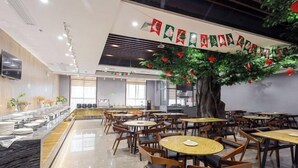 Un restaurant avec des tables et des chaises rondes, un espace buffet et un grand arbre artificiel décoré d’ornements rouges.