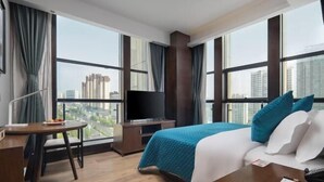 Une chambre d’hôtel équipée d’un lit, d’un bureau, d’une chaise, d’une télévision et de grandes fenêtres donnant sur la ville.