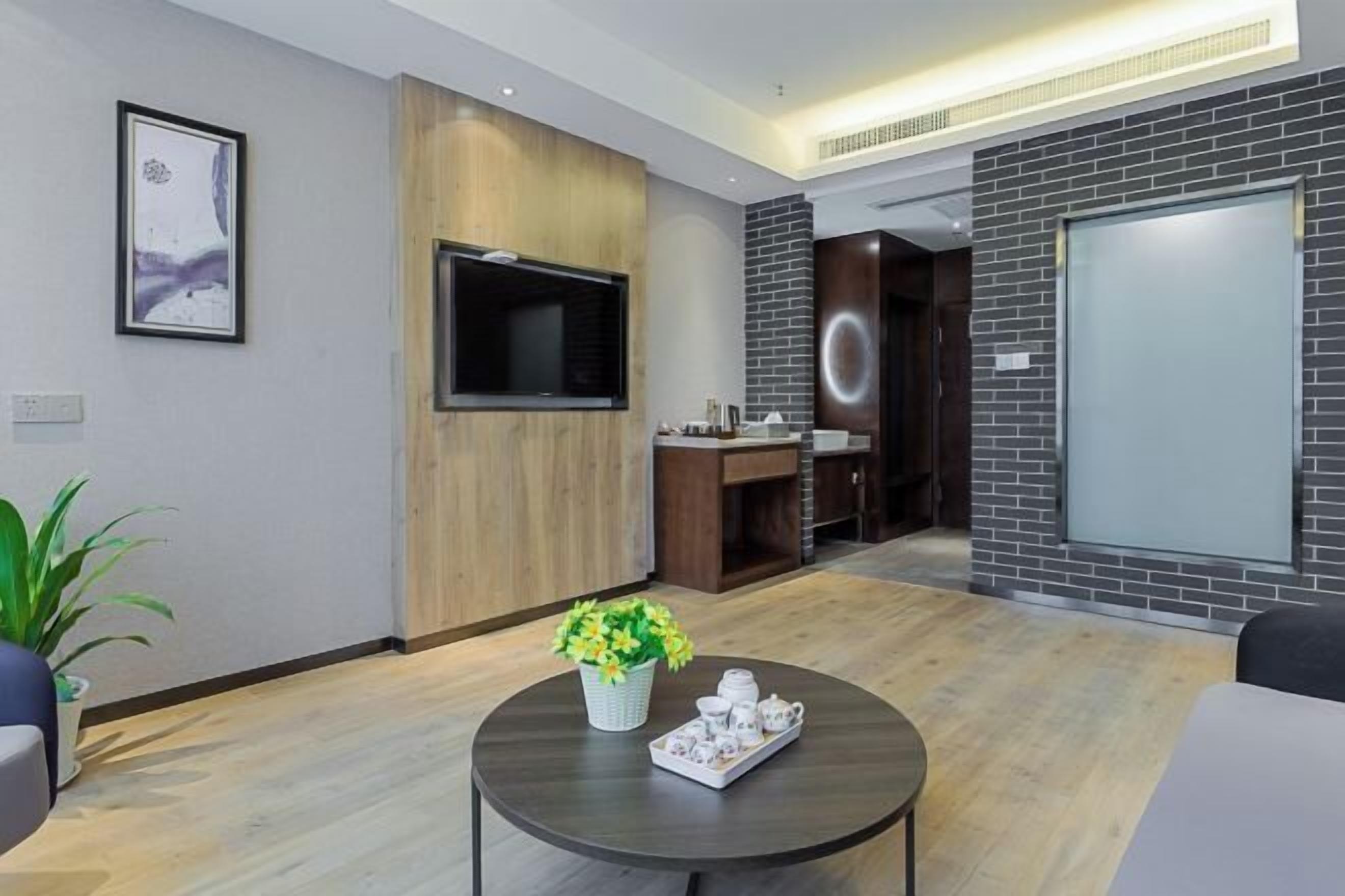 Una habitación de hotel moderna con una televisión de pantalla plana, un panel de madera decorativo, una mesa de centro redonda con una bandeja de tazas de té y una planta en maceta.