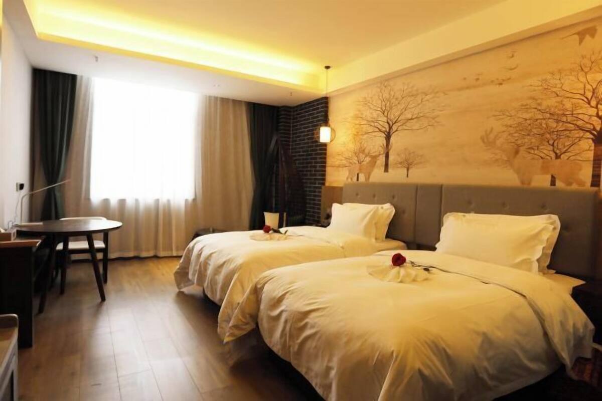 A hotel room with two beds, a dining table, a painting on the wall, and a window with curtains.