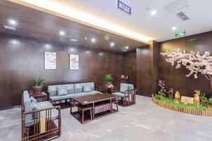 Un área de recepción con un conjunto de sofás, una mesa de centro y sillas, una pared decorativa con un arreglo floral, y un cartel que indica la ubicación.