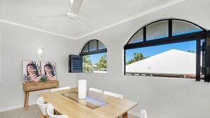 Un comedor con una mesa de madera, sillas blancas, un ventilador de techo y ventanas grandes con arcos.