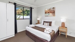 Habitación de hotel con cama, ventilador de techo, ventana con cortinas, un cuadro y mesita de noche con lámpara.