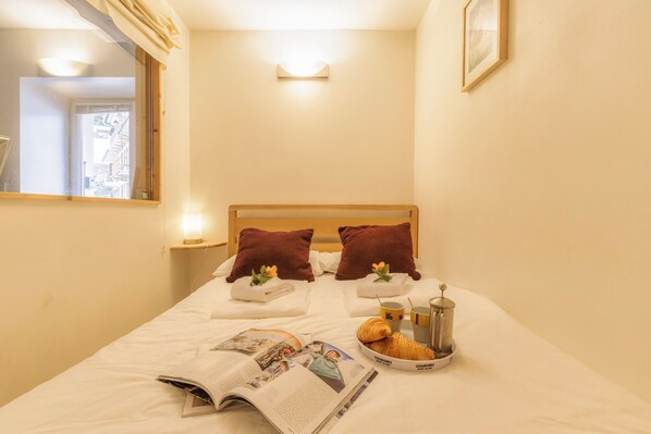 Een net opgemaakt bed met kussens, een tijdschrift, een croissant en een koffiezetapparaat.