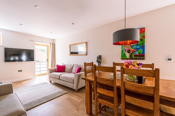 A modern living room with a sofa, a dining table with chairs, a flat-screen TV mounted on the wall, and a colorful painting above the table.