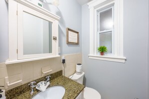 Ein Badezimmer mit einer Granit-Arbeitsplatte, einem weißen Waschbecken, einem Spiegel, einer Toilette und einem Fenster mit einer Pflanze.