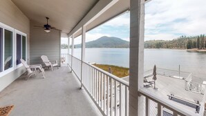 Un balcon couvert avec des balustrades blanches, deux fauteuils de jardin blancs, un ventilateur de plafond et une vue sur un lac, avec une montagne en arrière-plan.
